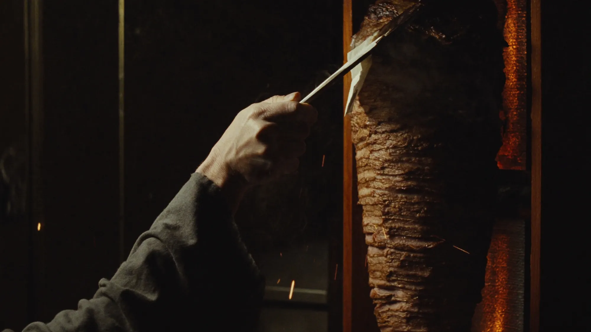 Eine Hand schneidet mit dem langen Messer eine dünne Scheibe vom Dönerspieß, dahinter glüht eine Wand aus Holzkohle.