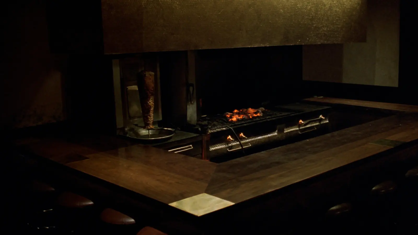 Der leere Gastraum am Abend: Eine Theke aus dunkler Eiche umläuft den Holzkohlegrill unter einer Haube aus Messing, links steht der Spieß.