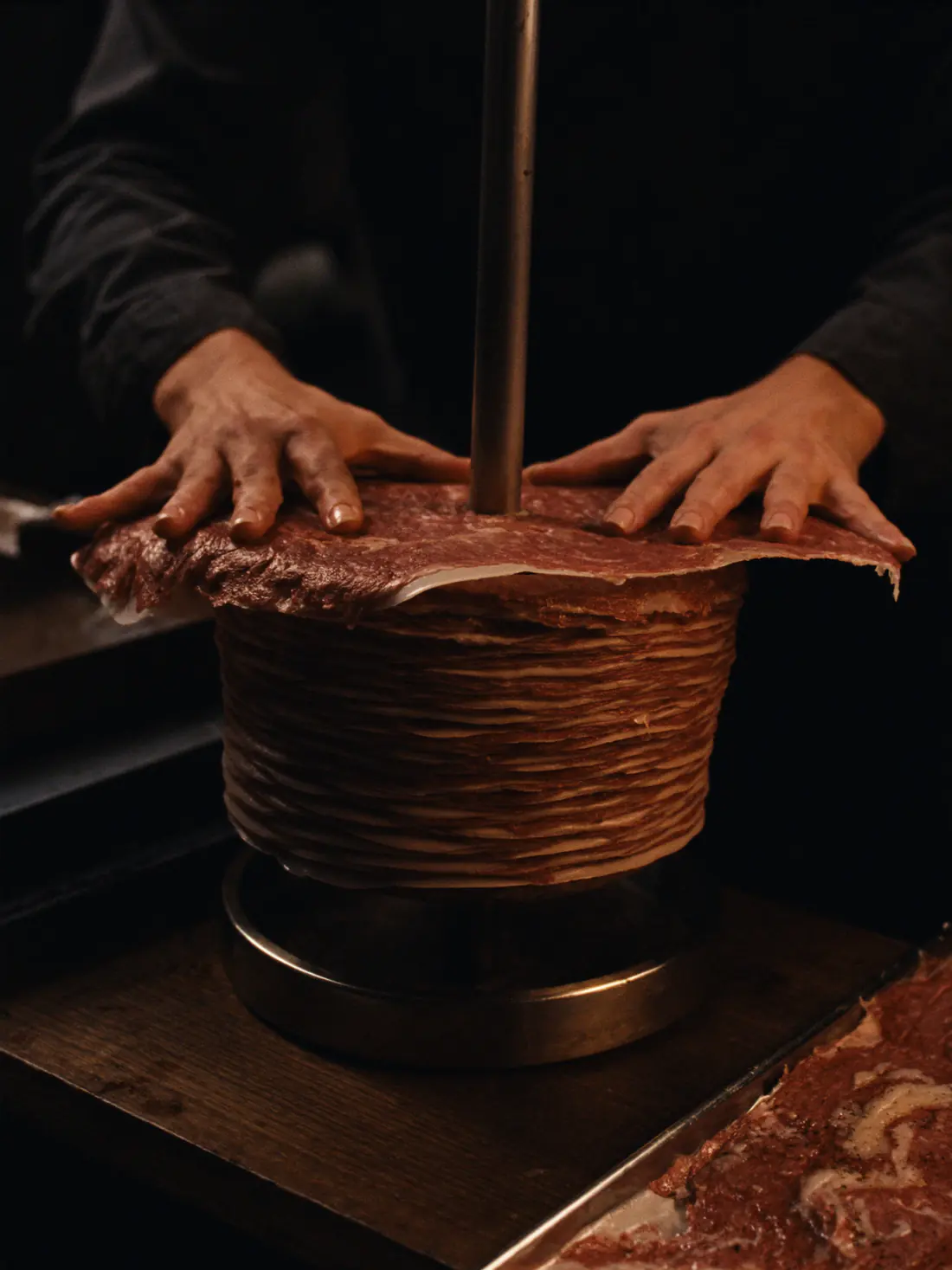 Zwei Hände drücken eine dünne Scheibe mariniertes Fleisch flach auf den Spieß. An der Seite sieht man die Lagen aus Fleisch und Fett, daneben liegt ein Blech mit weiteren Scheiben.